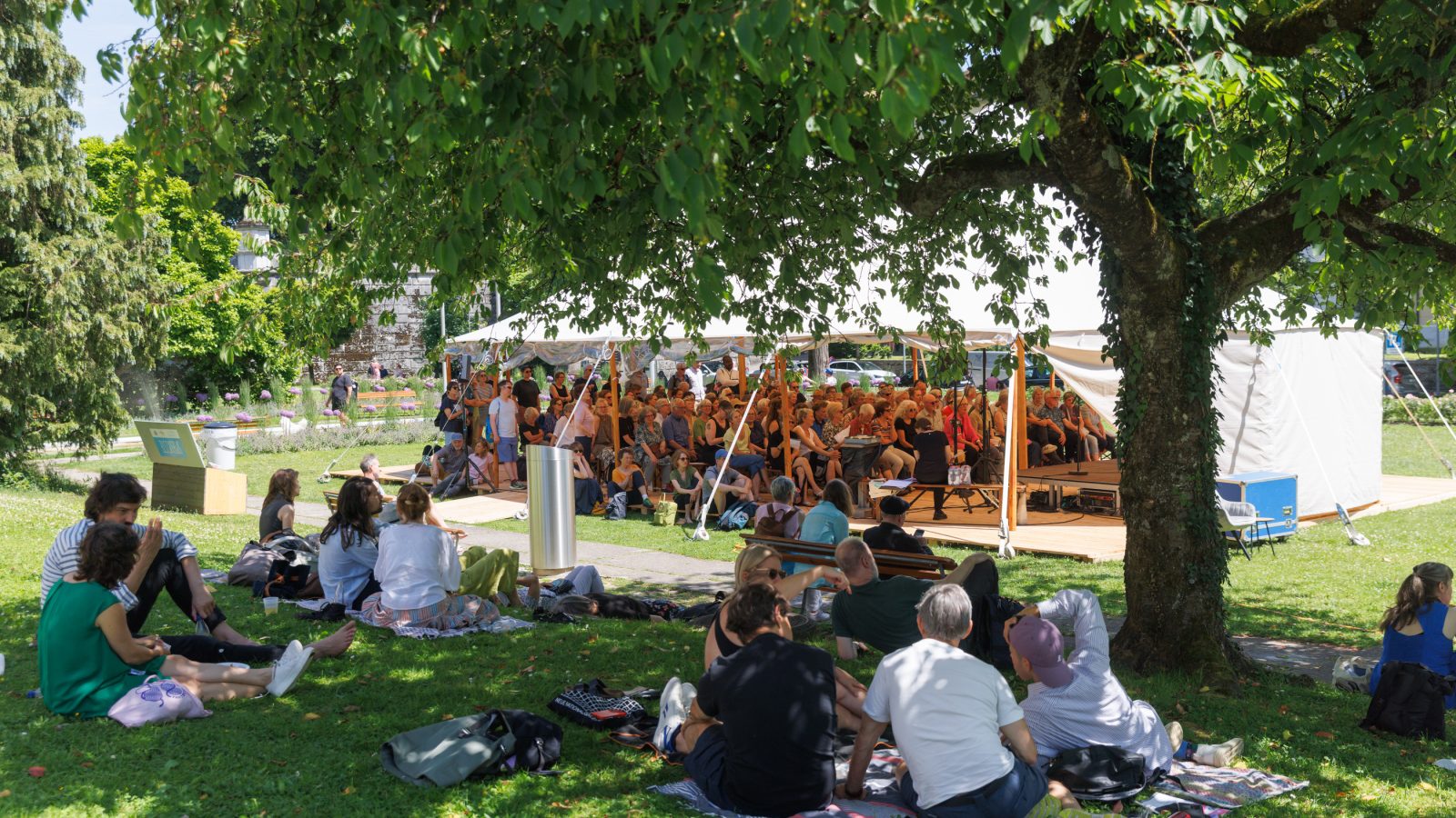 Il pubblico è seduto in una tenda all’interno di un parco verdeggiante, mentre davanti ad essa ci sono persone sedute sull’erba.