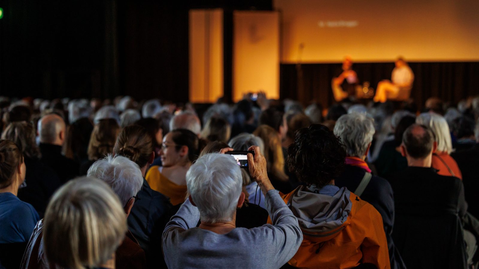 Evento con un folto pubblico; sul palco sono sedute due persone.